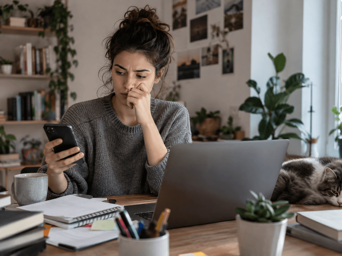 A stressed-looking woman at her desk checks her phone instead of the open laptop in front of her.
